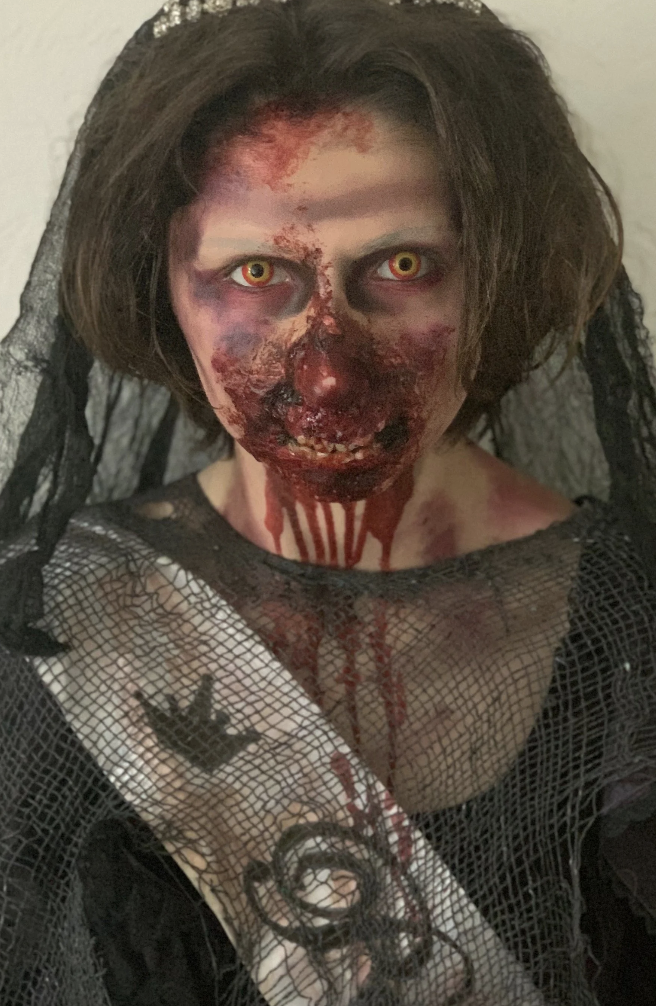 Person dressed as a zombie or monster with blood, yellow eyes, and torn clothing, wearing black lace and a headpiece with black fabric.
