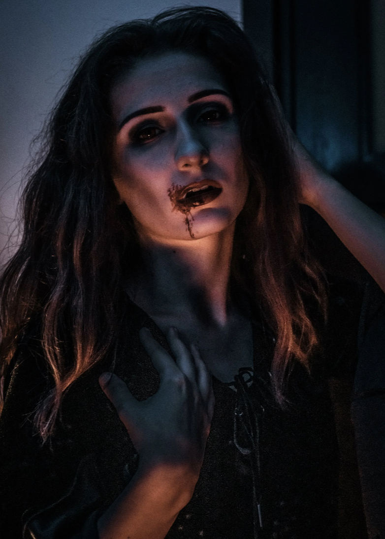 A woman with dark, wavy hair and dark makeup, including dark lipstick and eye makeup, has a wound on her lip with blood drips. She is holding her neck with one hand and touching her head with the other, in a dimly lit room with blue and purple lighti