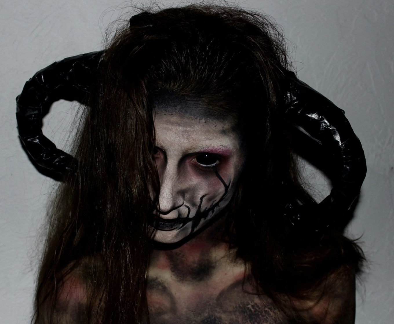 Person with long hair wearing makeup designed to look like a skull, with black horns on their head, standing against a plain wall.