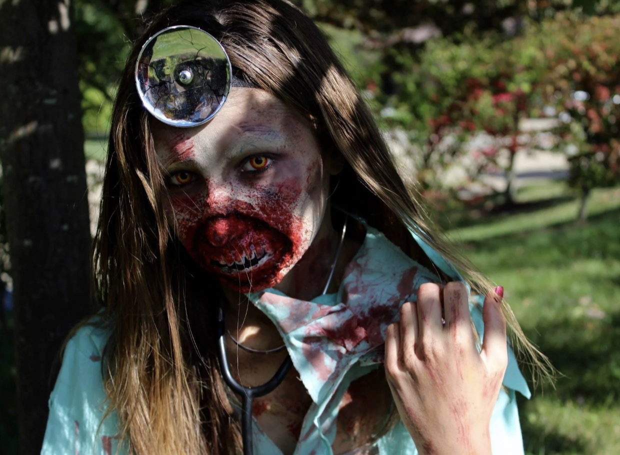 Person wearing a zombie costume with blood and makeup, head tilted, outdoors in sunlight, with trees and foliage in background.