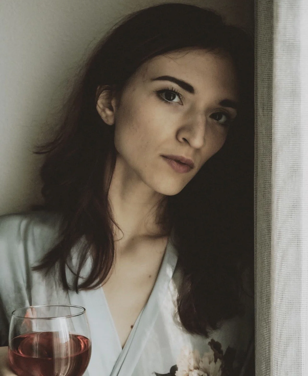 A woman with dark hair and light skin looking out a window, holding a glass of red wine.