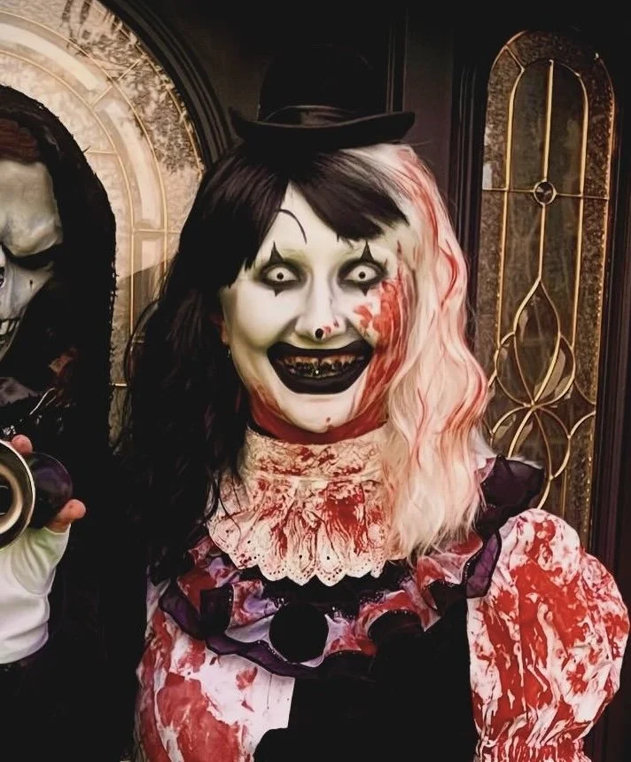 Person dressed in a creepy clown costume with blood-spattered clothing, white face makeup with black eyes, and exaggerated makeup, standing indoors near a golden decorative door.