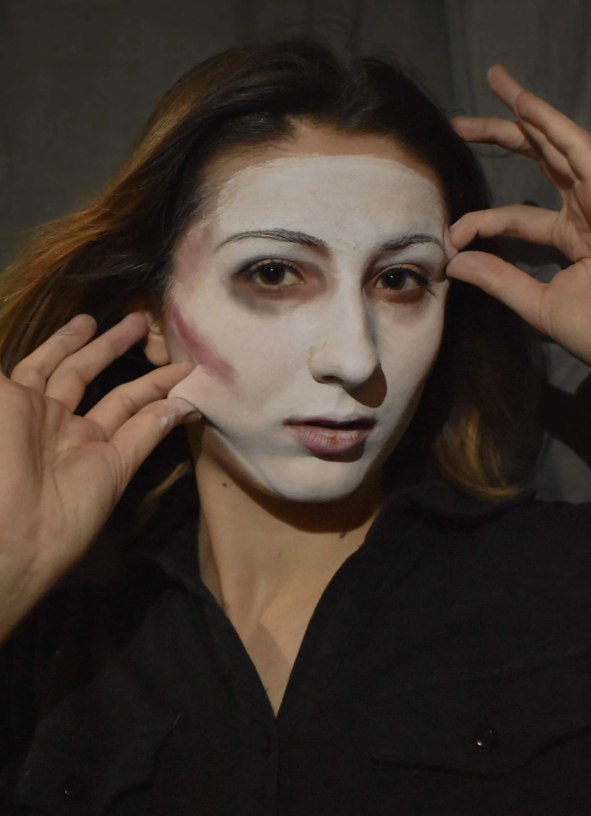 A woman with white face paint, dark eye makeup, and dark lipstick looking at the camera with her hands near her face.