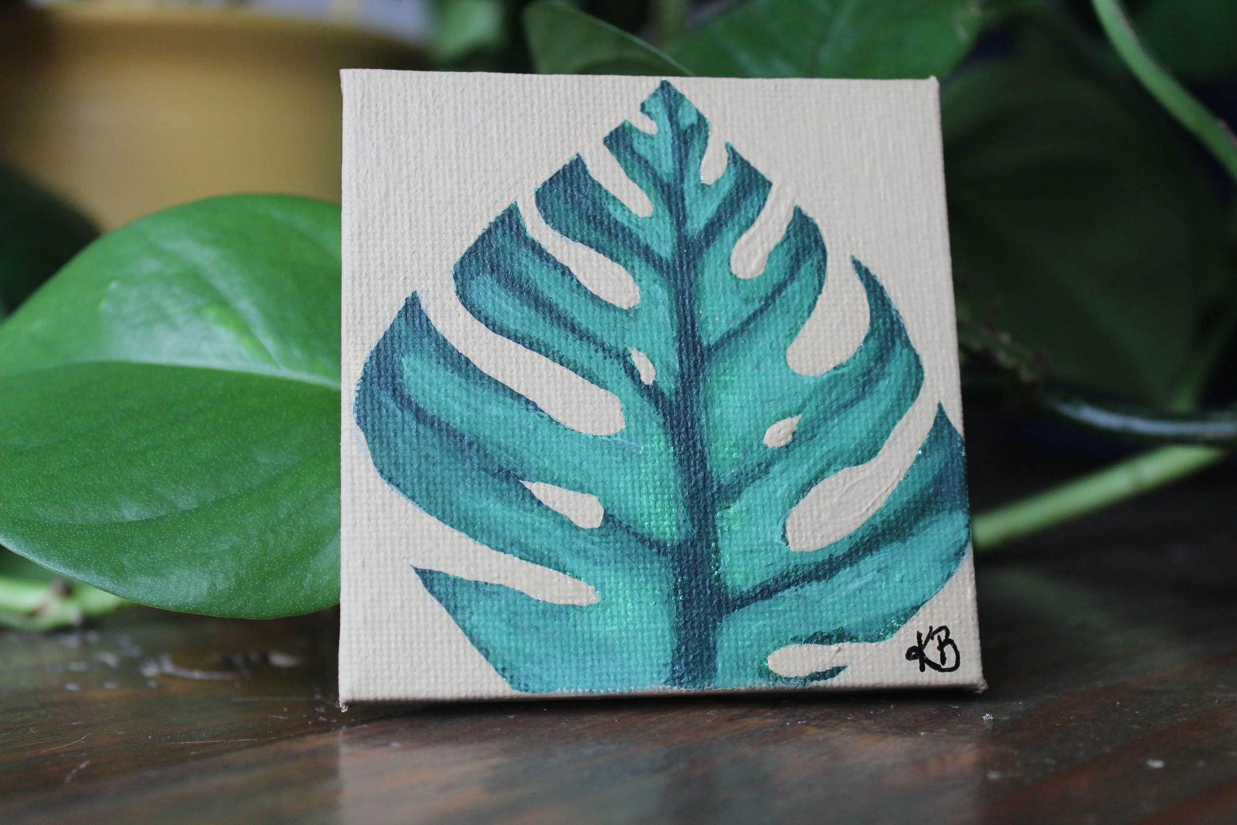 A small canvas painting of a blue-green Monstera leaf on a wooden surface, with green leaves in the background.