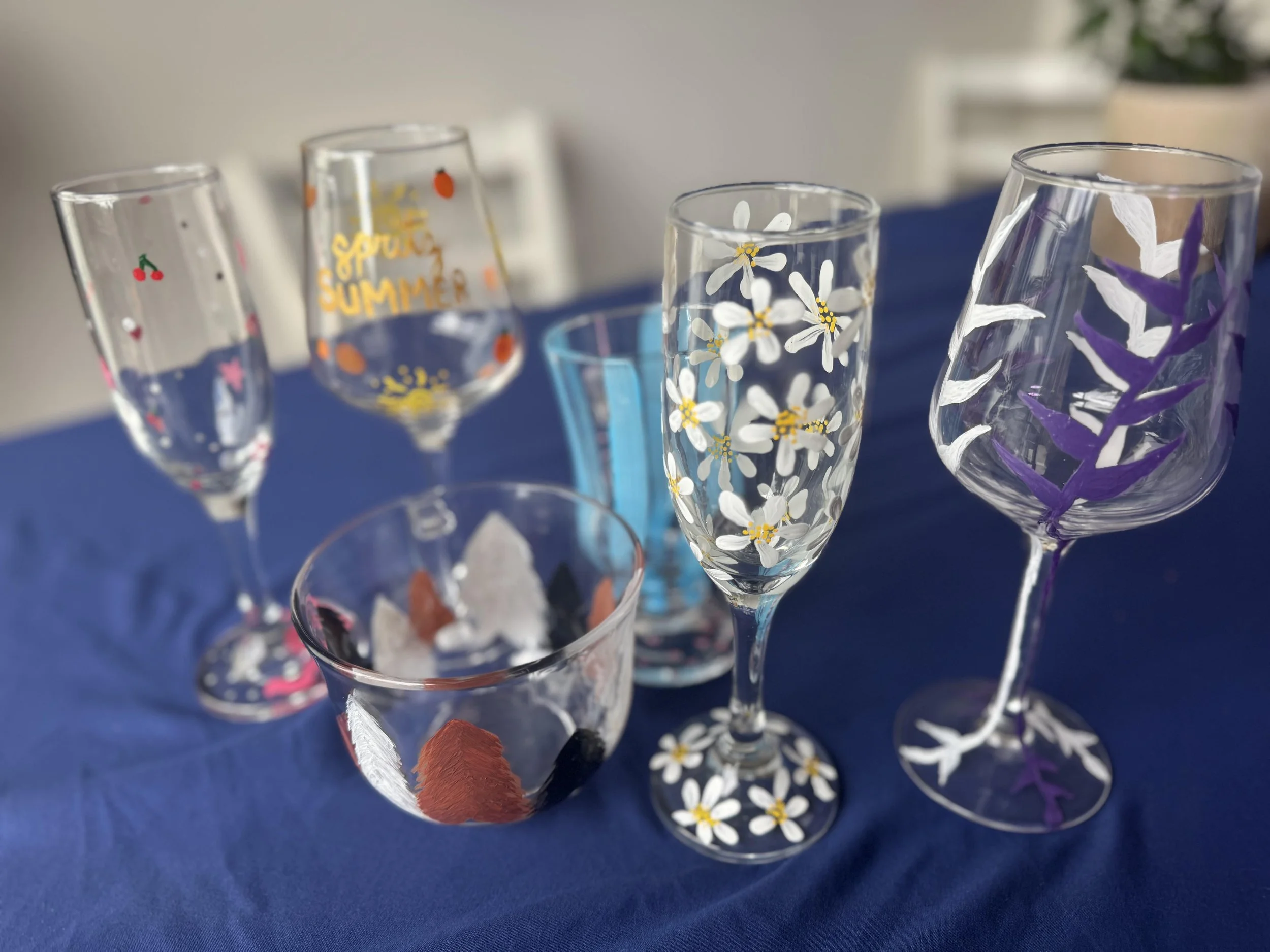 Six decorated glasses with various floral and summer-themed paintings on a blue tablecloth.