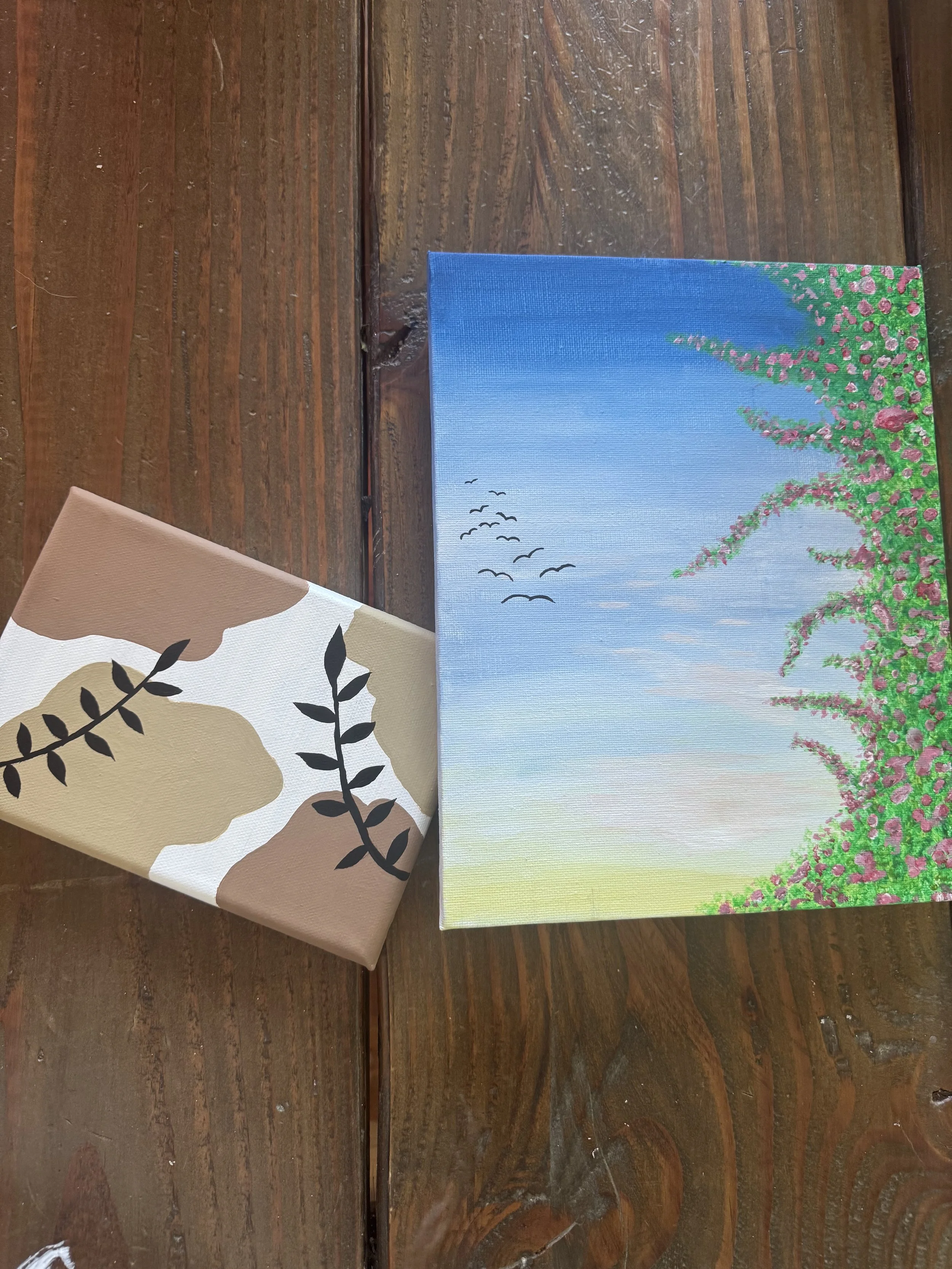 A small canvas painting of a nature scene with a blue sky, pink flowers, and green foliage with birds flying in the sky, placed on a wooden table.
