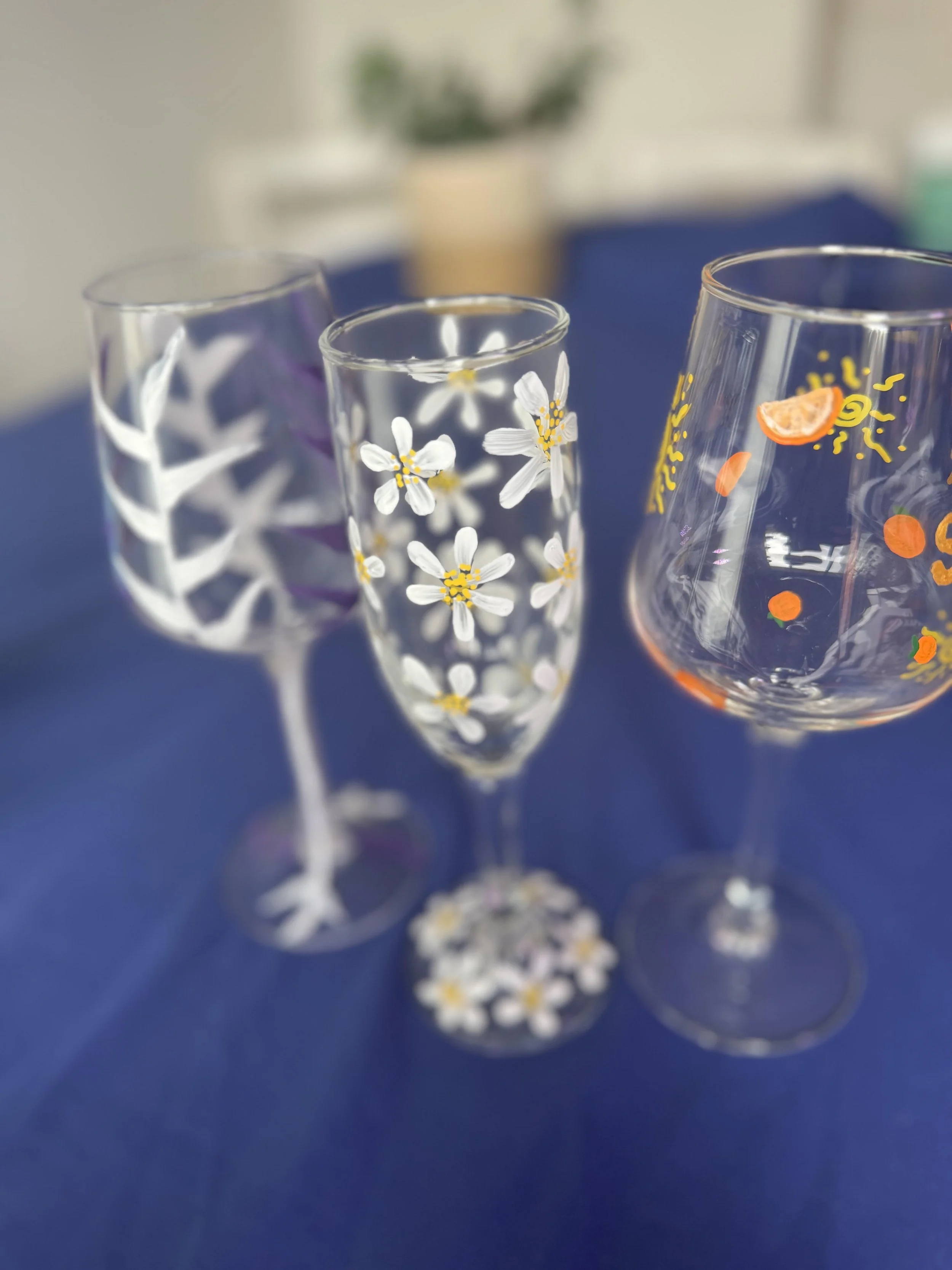 Three decorated glassware items on a blue tablecloth, including a glass with white and yellow daisy patterns, a glass with a purple and white leafy pattern, and a large glass with colorful fruit and sun designs.