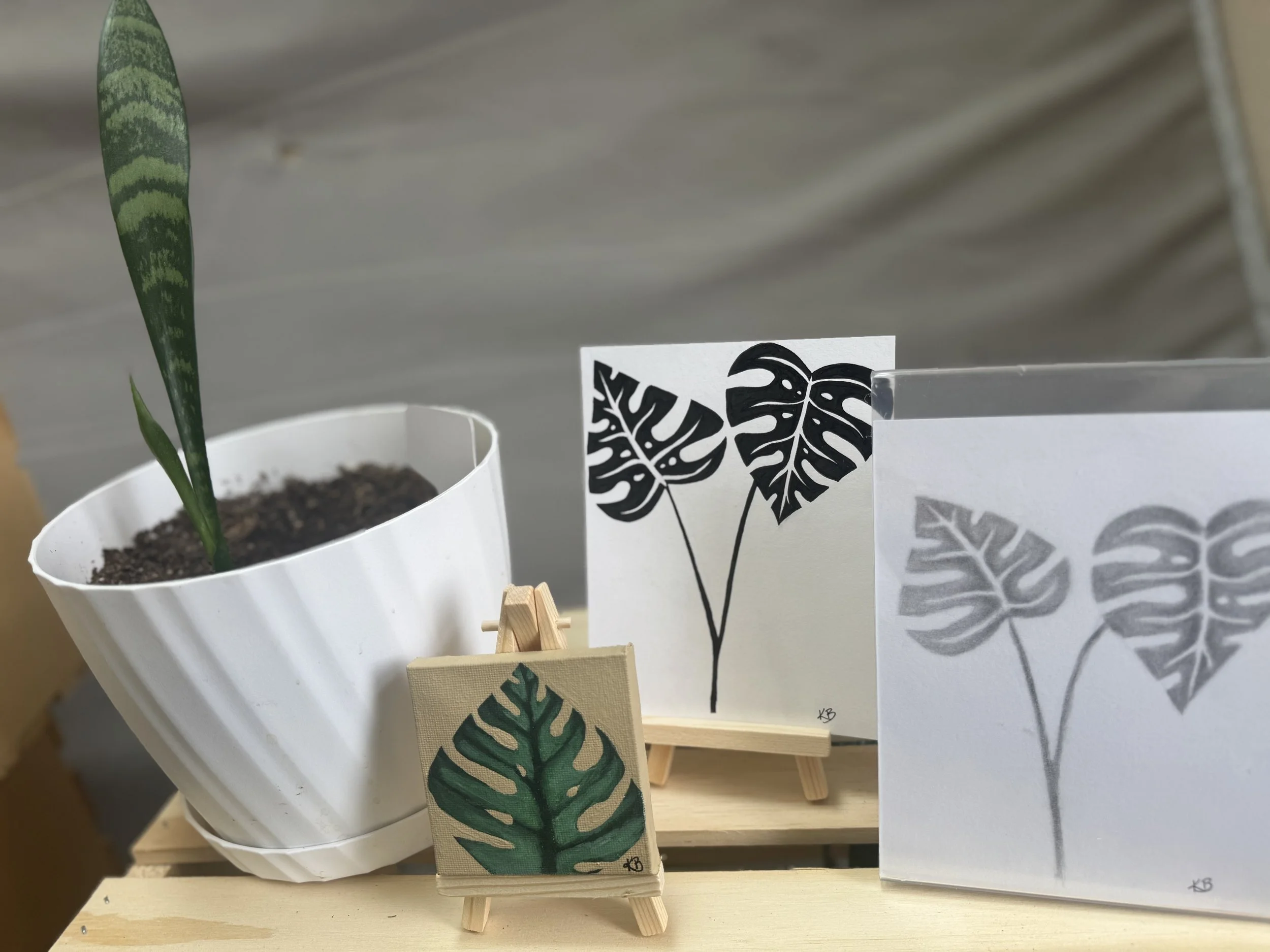 A potted plant with a tall, green, variegated leaf next to art prints of the Monstera leaf pattern, one in color and two in black and white, displayed on small easels on a wooden surface.