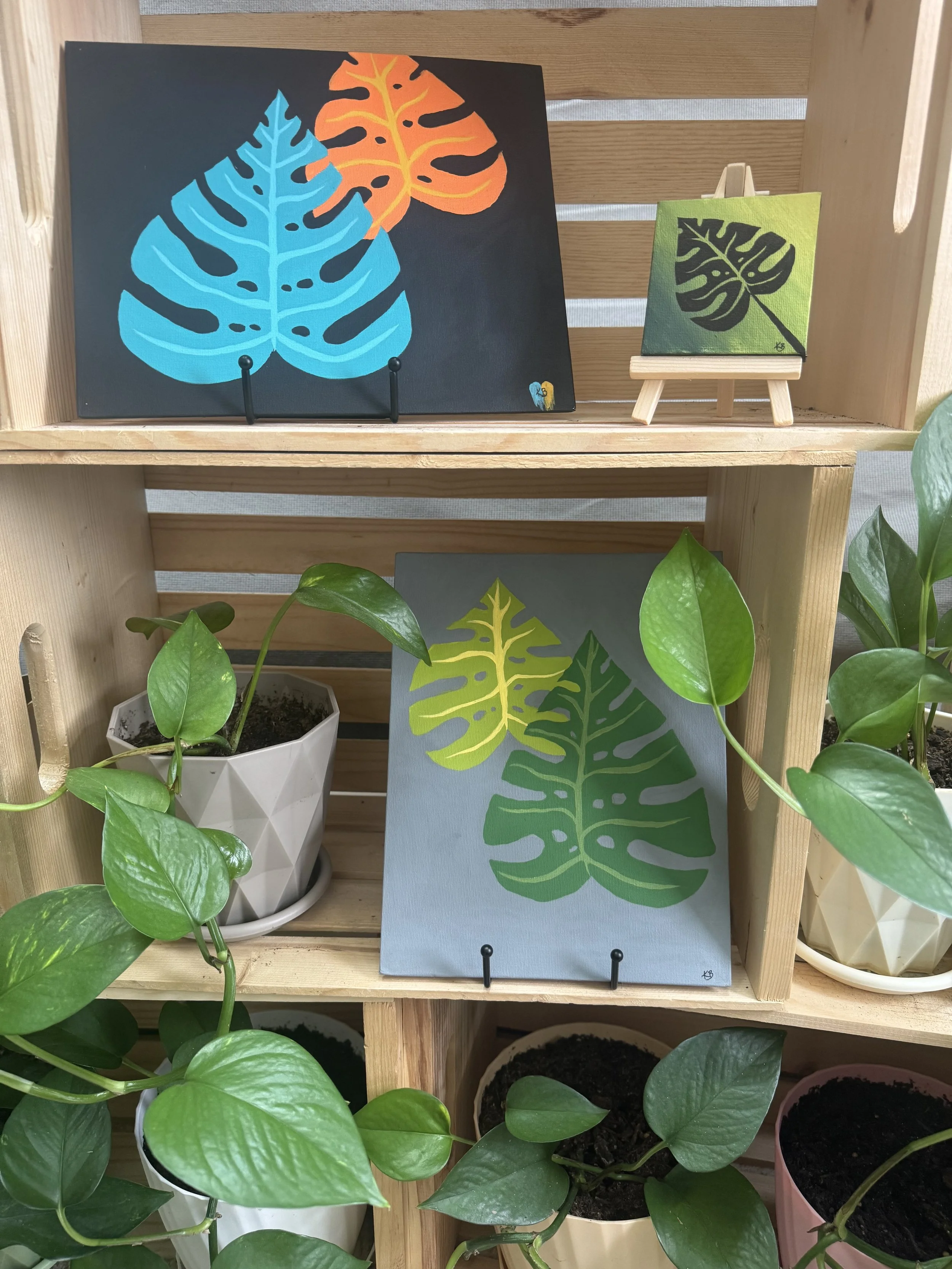 Displays three paintings of monstera leaf designs on wooden shelves, surrounded by potted green plants.