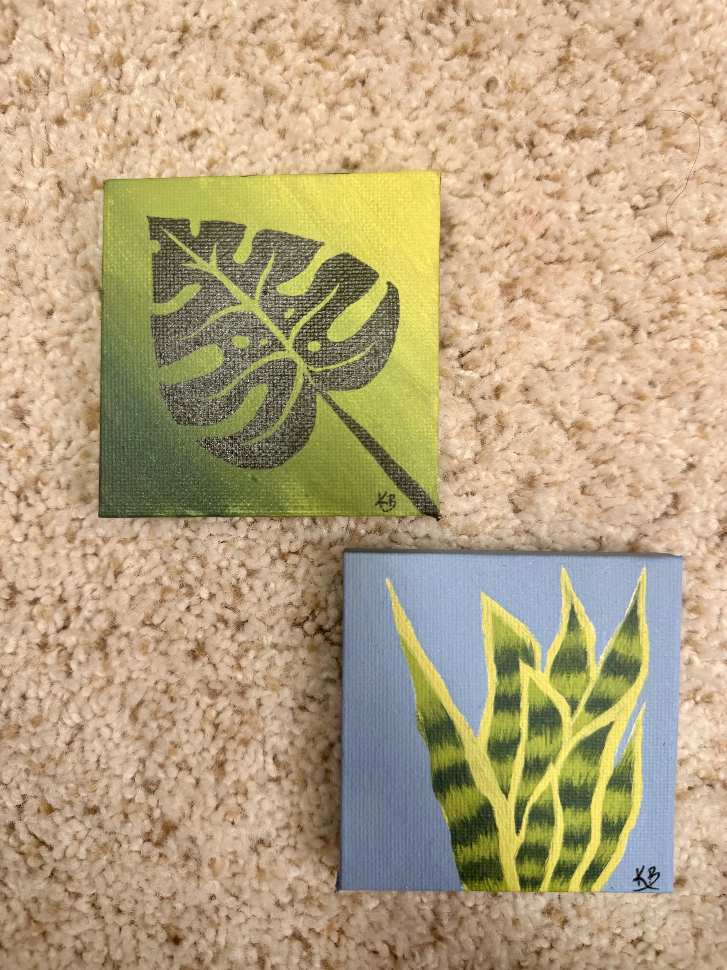 Two small square paintings of leaves on a beige carpet. The top painting depicts a green Monstera leaf with black detailing, and the bottom painting features a yellow and green striped leaf on a blue background.