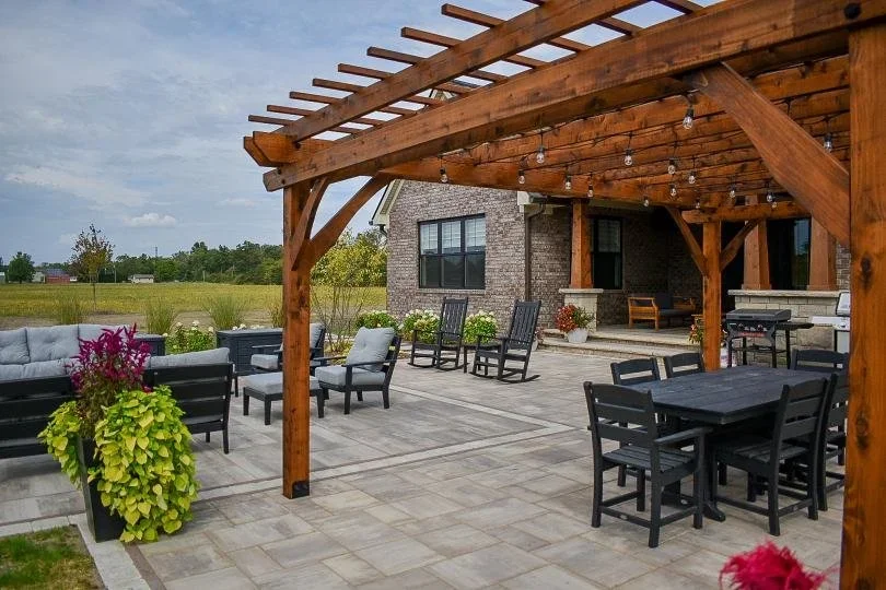 Outdoor patio area with a wooden pergola, outdoor furniture including chairs, sofas, a dining table, and potted plants, with a grassy field and house in the background.