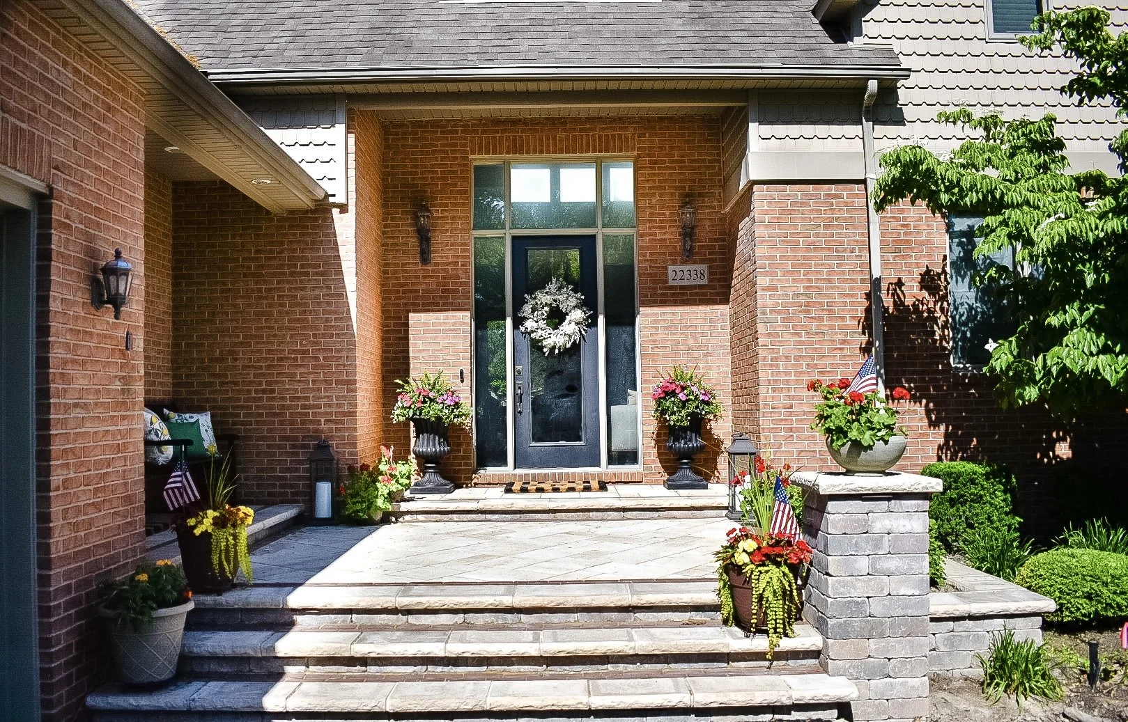 Front porch of a brick house decorated with potted flowers, American flags, and a white wreath on the door.