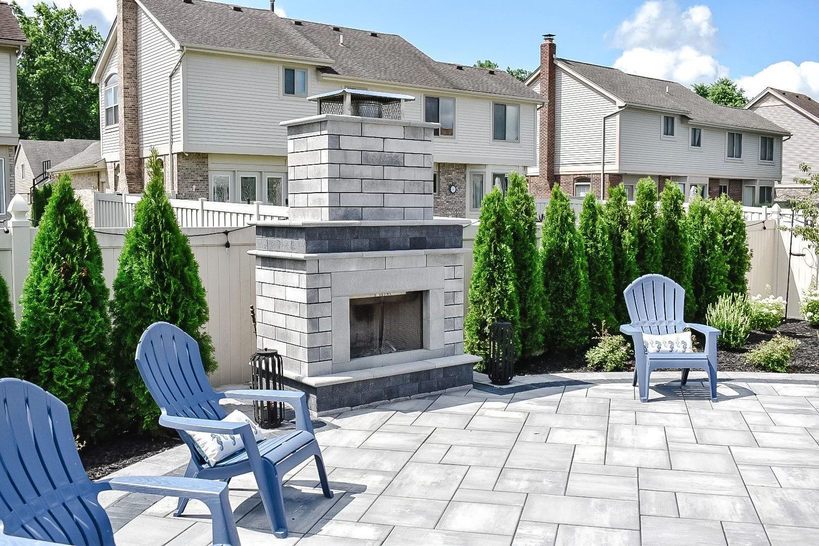 A backyard patio with a built-in stone fireplace, surrounded by a white fence, green shrubs, and outdoor chairs.