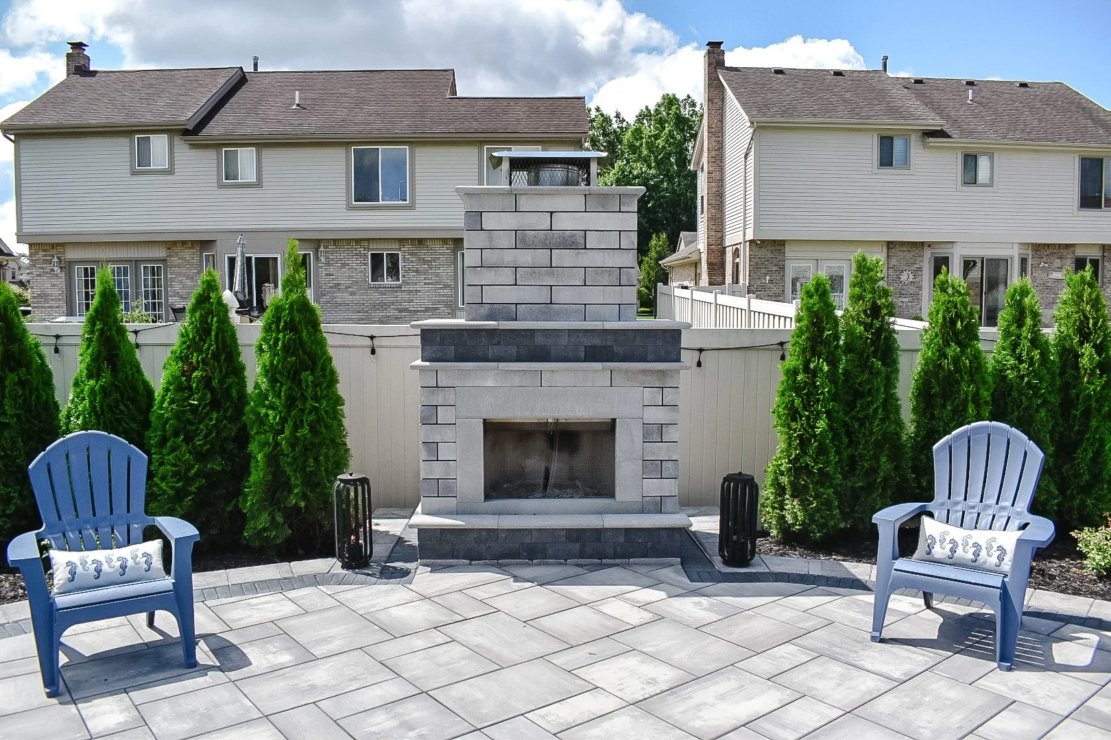 Backyard patio with a stone fireplace, two blue chairs with seahorse pillows, green shrubbery, and neighboring houses in the background.