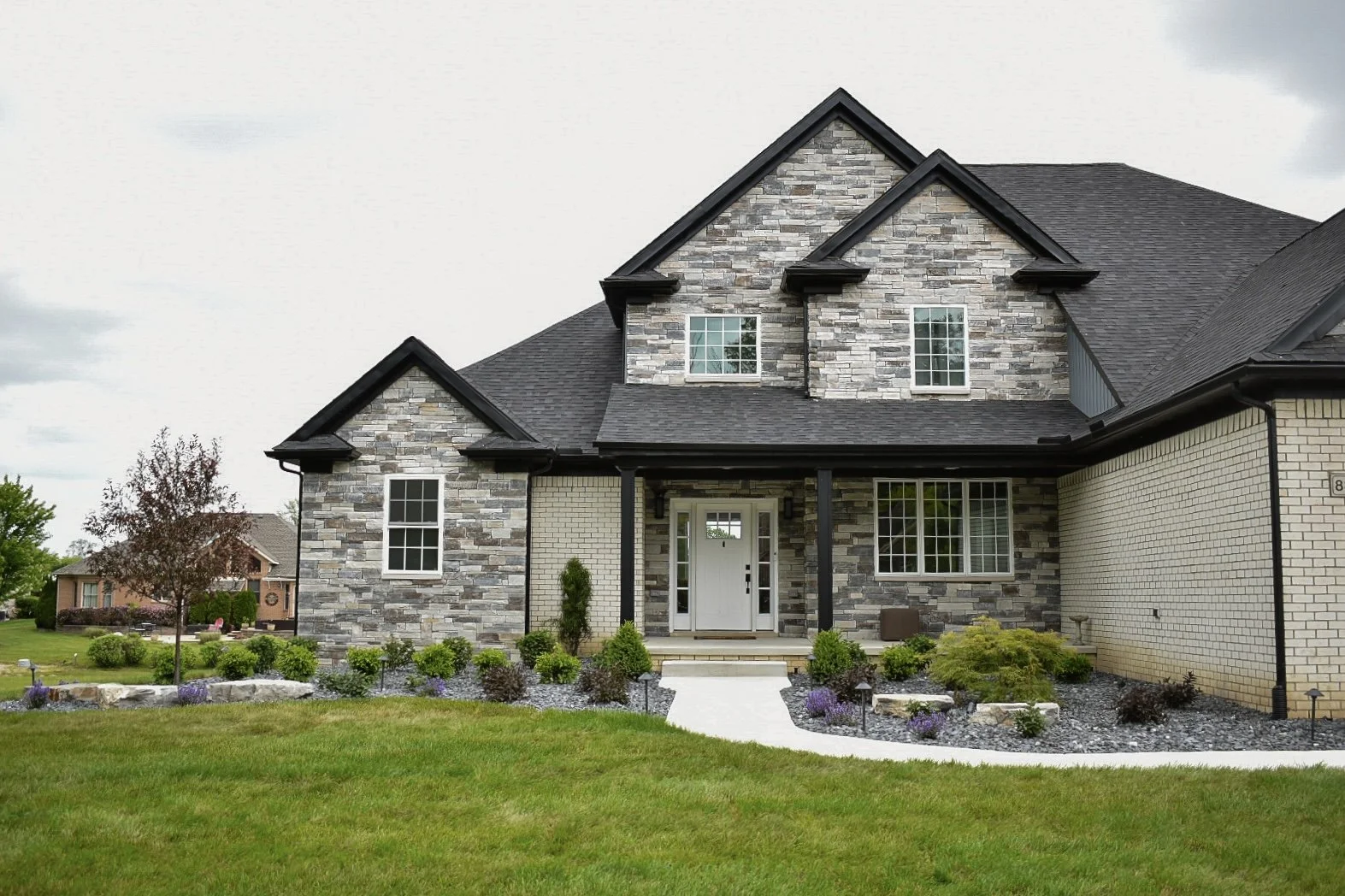A modern two-story house with a stone and brick exterior and black roof, surrounded by a well-manicured lawn and landscaping.