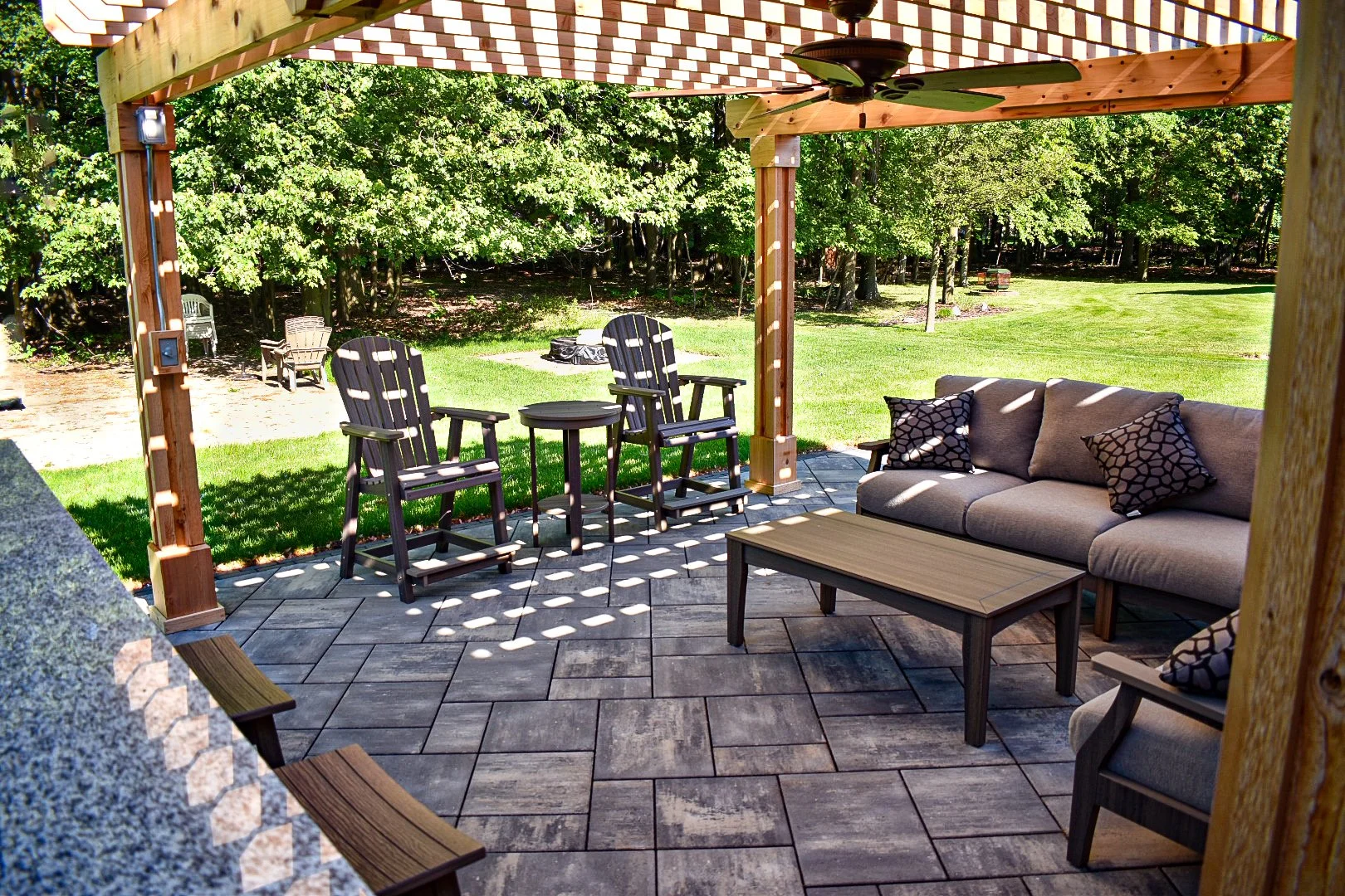 Outdoor patio area with a gray couch, black pillows, a wooden coffee table, and wooden Adirondack chairs on a stone paver floor, shaded by a pergola, overlooking a lush green backyard with trees and grass.