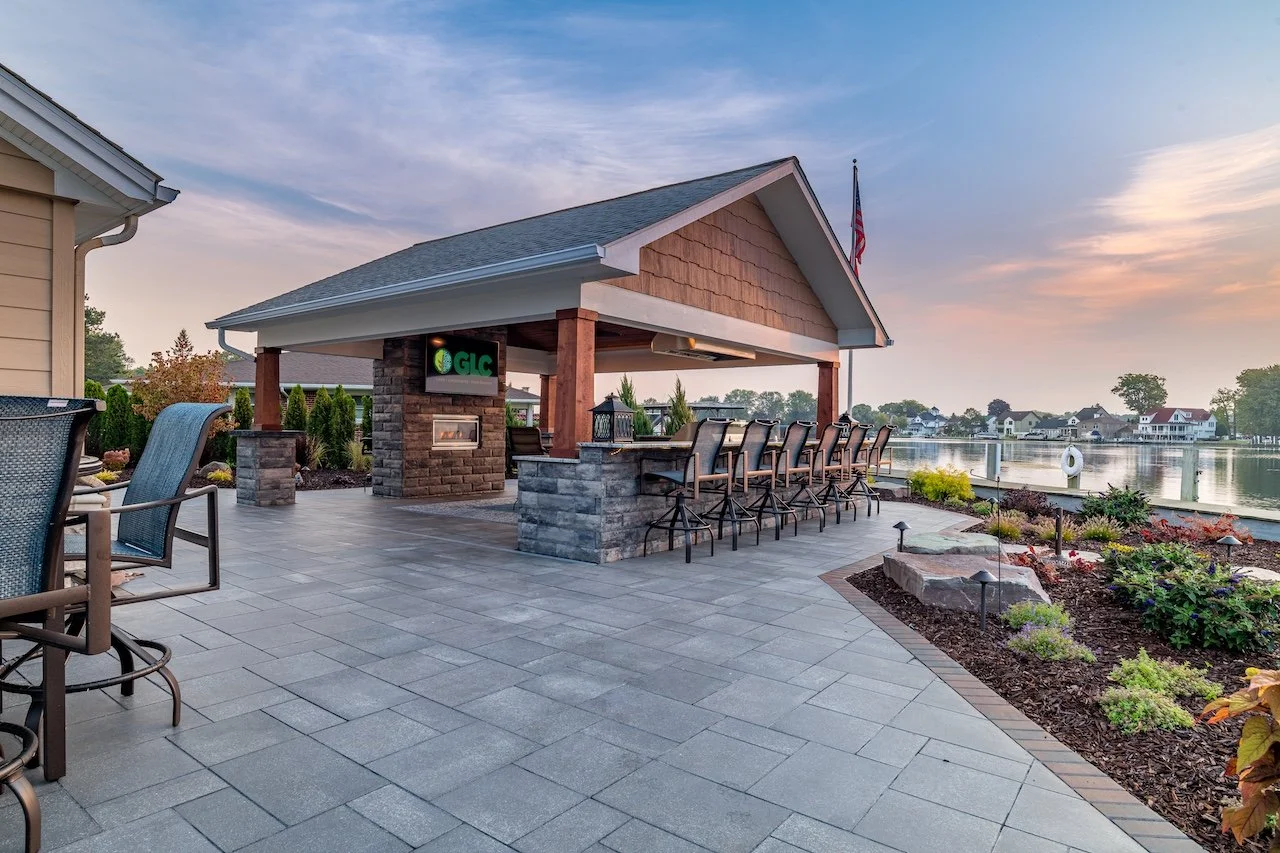 Outdoor patio by a lake with bar seating, flower beds, and a small pavilion with an American flag, during sunset.