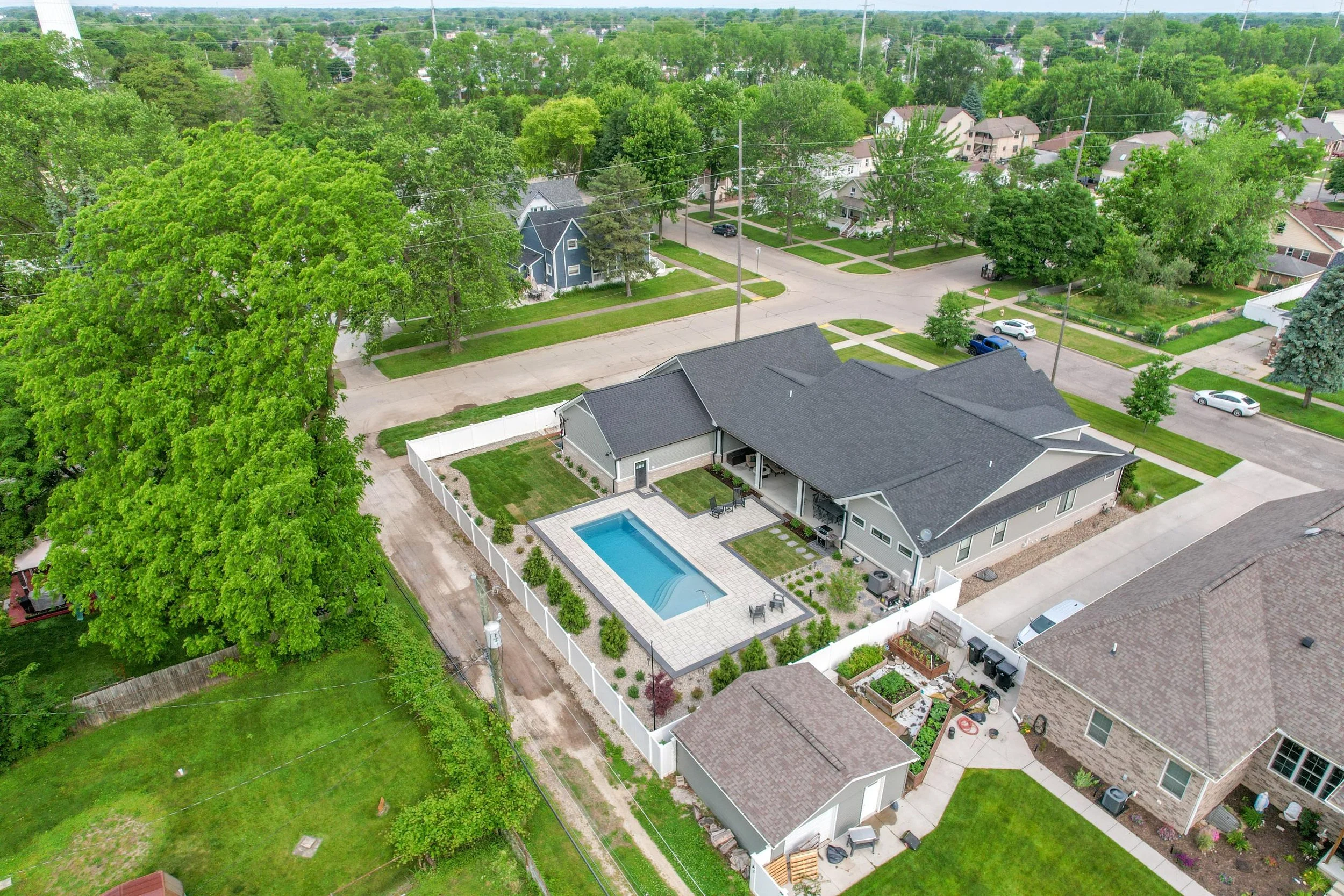 Aerial view of a suburban neighborhood during spring with green trees, houses, cars parked along the streets, and a backyard with a swimming pool surrounded by a white fence.