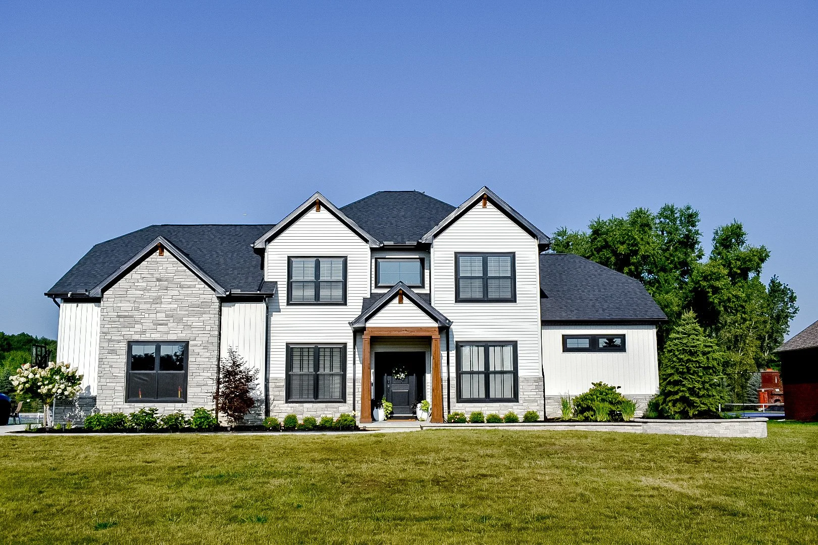 A modern two-story house with white and gray exterior, black door with a wreath, landscaped front yard, and blue sky.