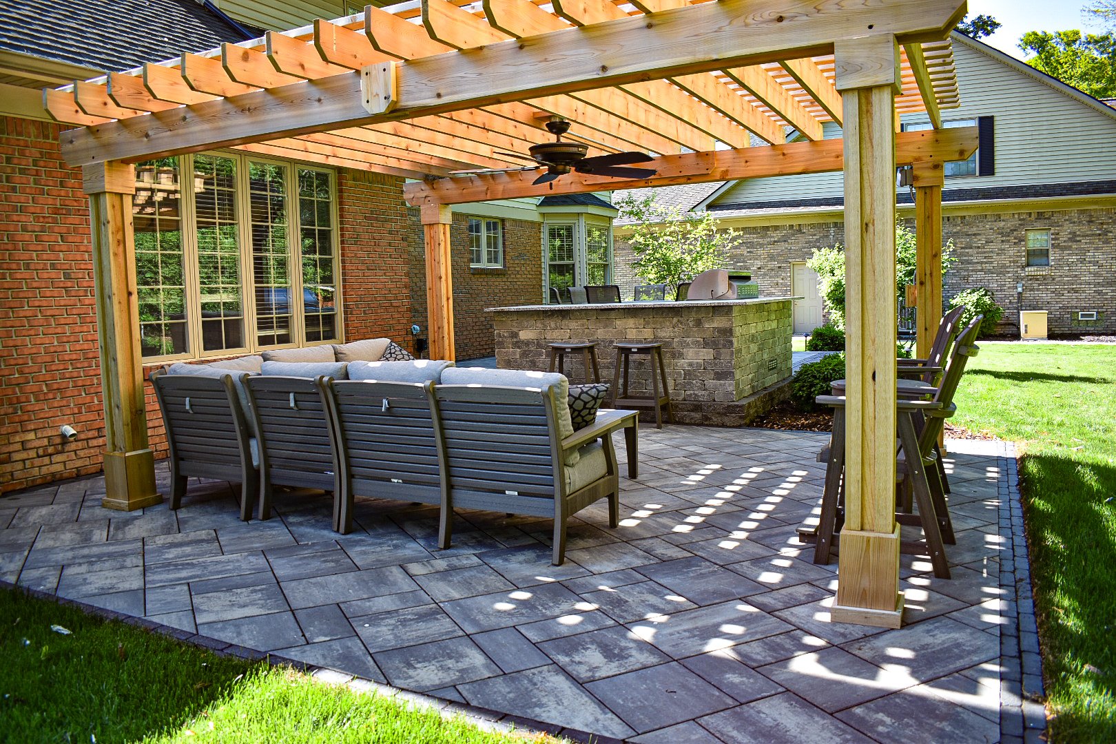 A backyard patio with a wooden pergola, outdoor seating, a brick bar with bar stools, and a grassy lawn in the background.