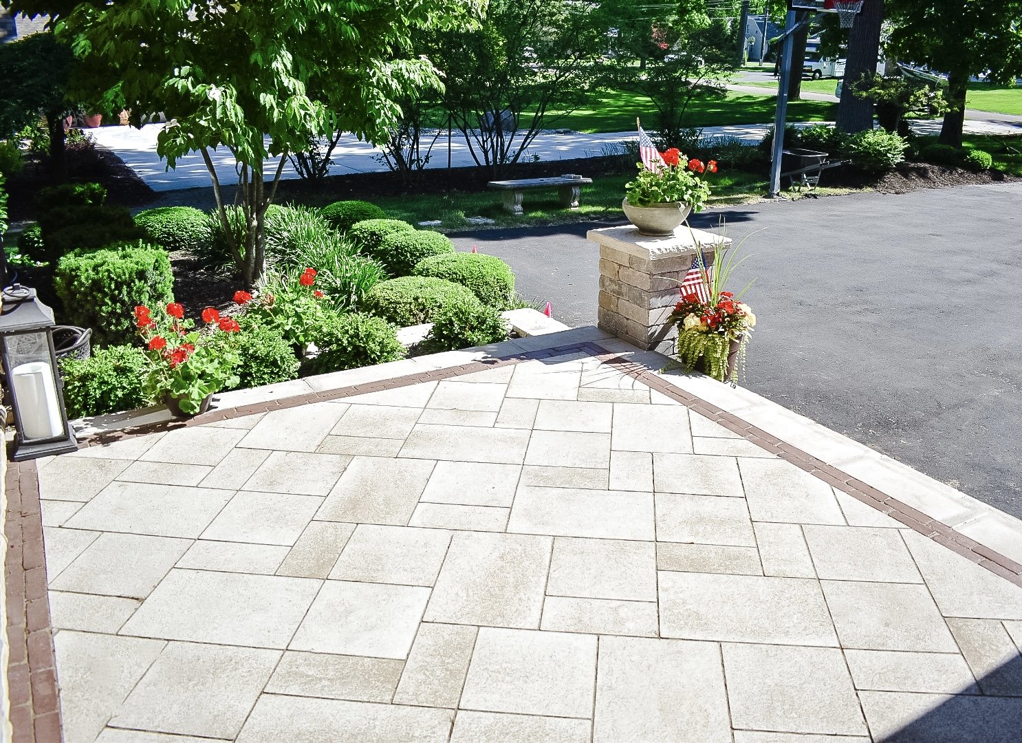 A front porch with beige stone tiles, two flower pots with red and white flowers and American flags, and a brick pillar. There is a parking lot and a green lawn with trees and bushes in the background.