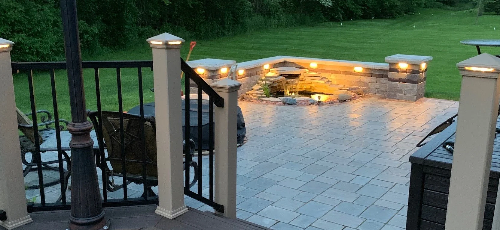 A cozy outdoor dining area under a patio with string lights in a yard during evening, with chairs around a table set for a meal and surrounded by potted plants and greenery — GLC Lawn & Landscape