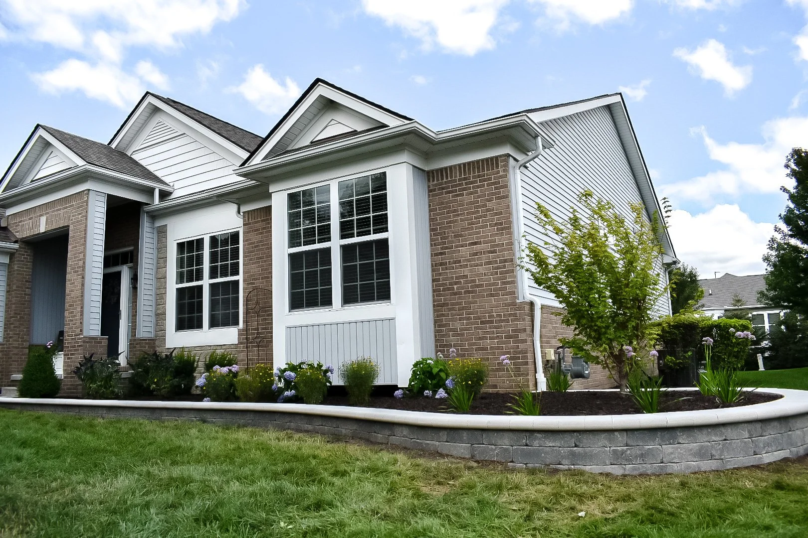 A modern house with brick and white siding exterior, large front windows, and a small garden with flowers and bushes in the front yard.