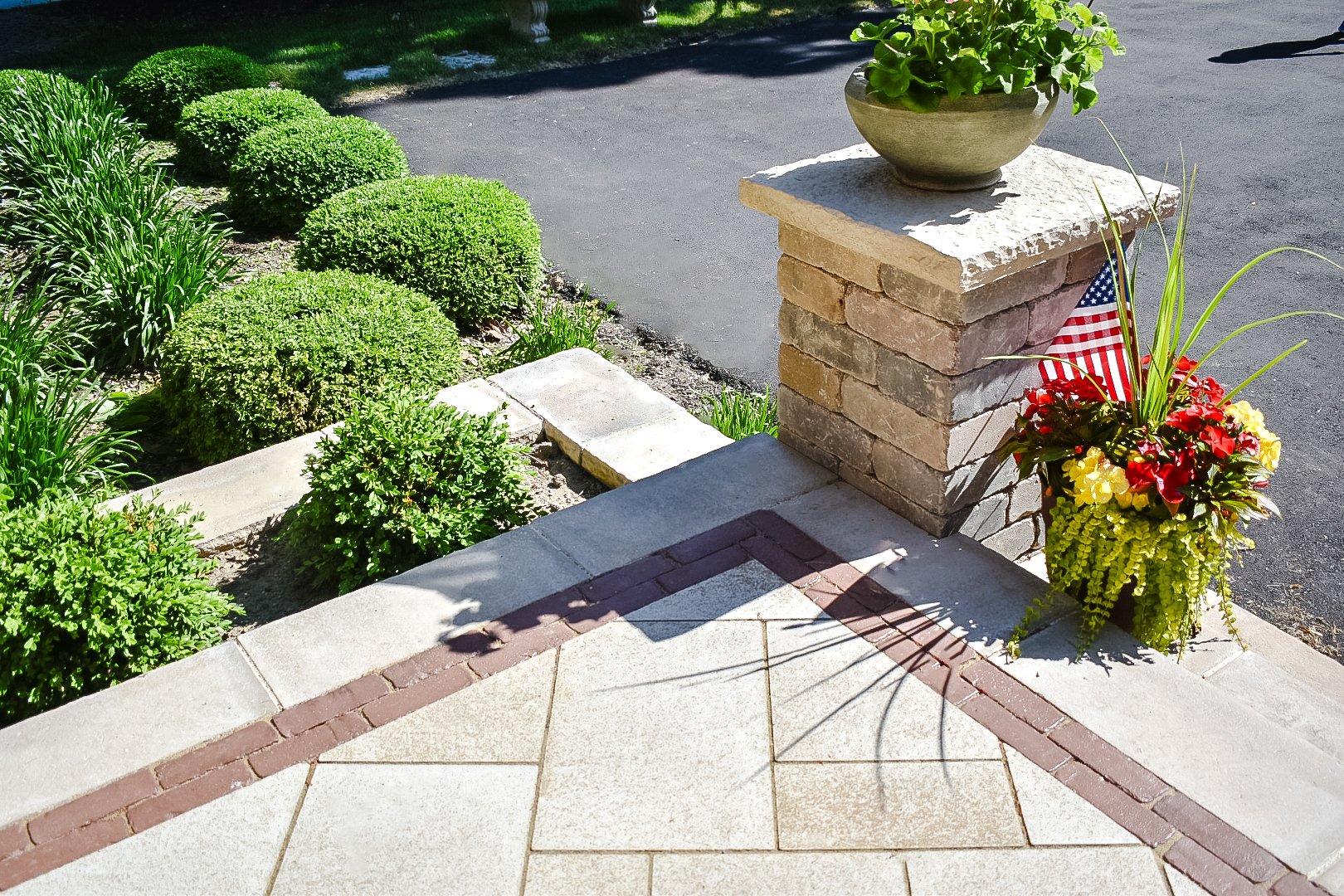 Decorative concrete and brick porch with a brick pillar, a potted plant on top, a flower arrangement with an American flag, and neatly trimmed bushes in the garden.