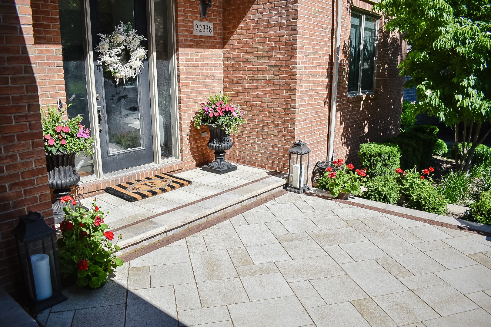The front porch of a brick house decorated with potted flowers, a festive wreath on the door, and outdoor lighting, with a landscaped garden on the side.