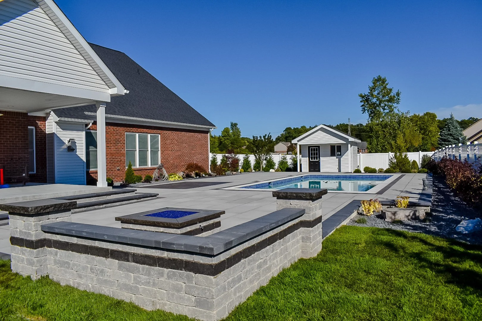 Backyard with a swimming pool, a small shed, landscaped garden beds, a brick house, and a white privacy fence under a clear blue sky.