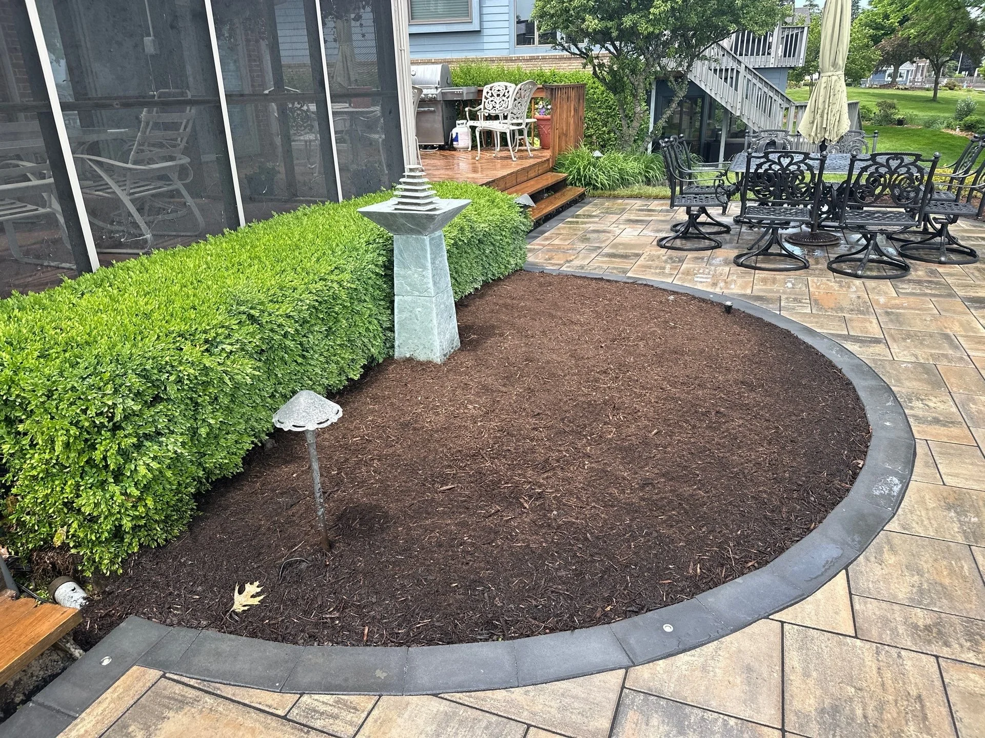 A backyard patio with a round black metal table and six matching chairs, a large beige patio umbrella, a wooden deck with a white metal patio set, lush green bushes, a tree, and house stairs made of wood leading to an upstairs deck. There is a stone 