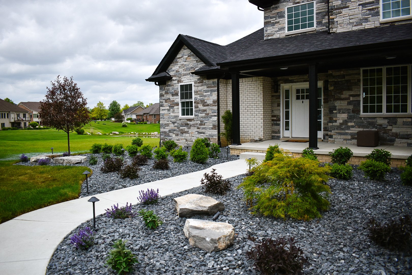 Front view of a house with a porch and landscaped yard, featuring dark gray composite siding, stone accents, and white-framed windows. The yard has a concrete walkway, decorative rocks, small bushes, and landscaping lights under an overcast sky.