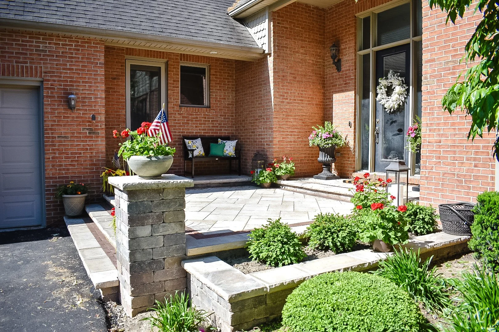 Front porch of a brick house decorated with flower pots, American flags, and outdoor furniture, with a landscaped garden and steps leading up to the door.