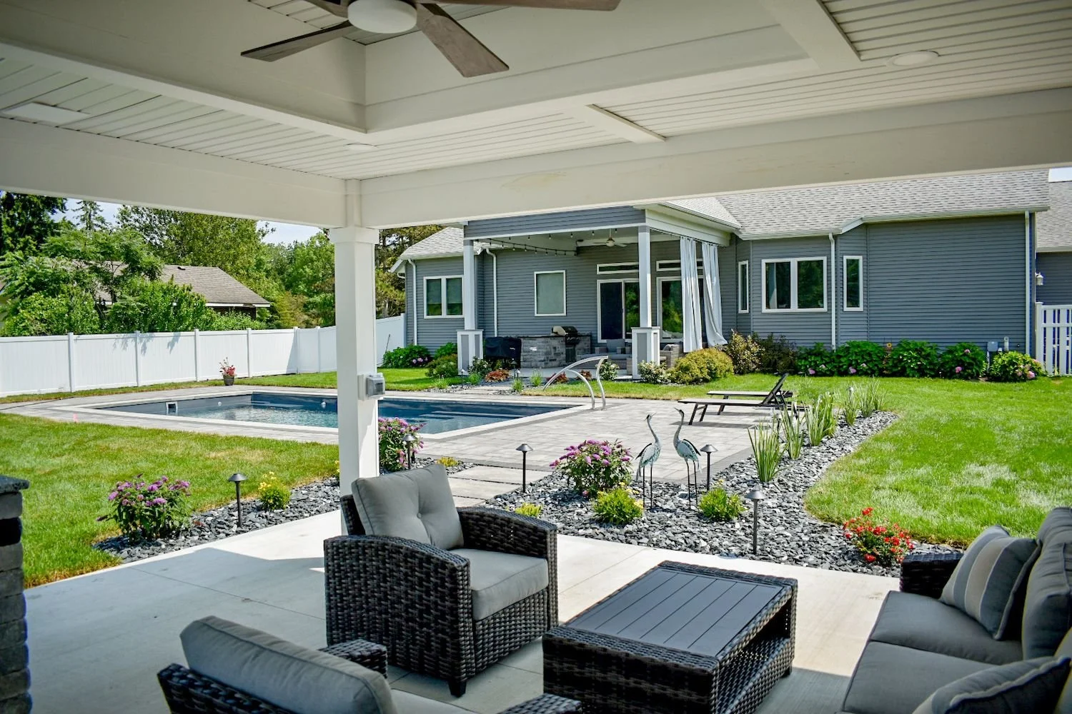 View of a backyard patio with outdoor furniture, overlooking an in-ground swimming pool and a well-maintained lawn with garden decorations, surrounded by a white fence and a blue house in the background.