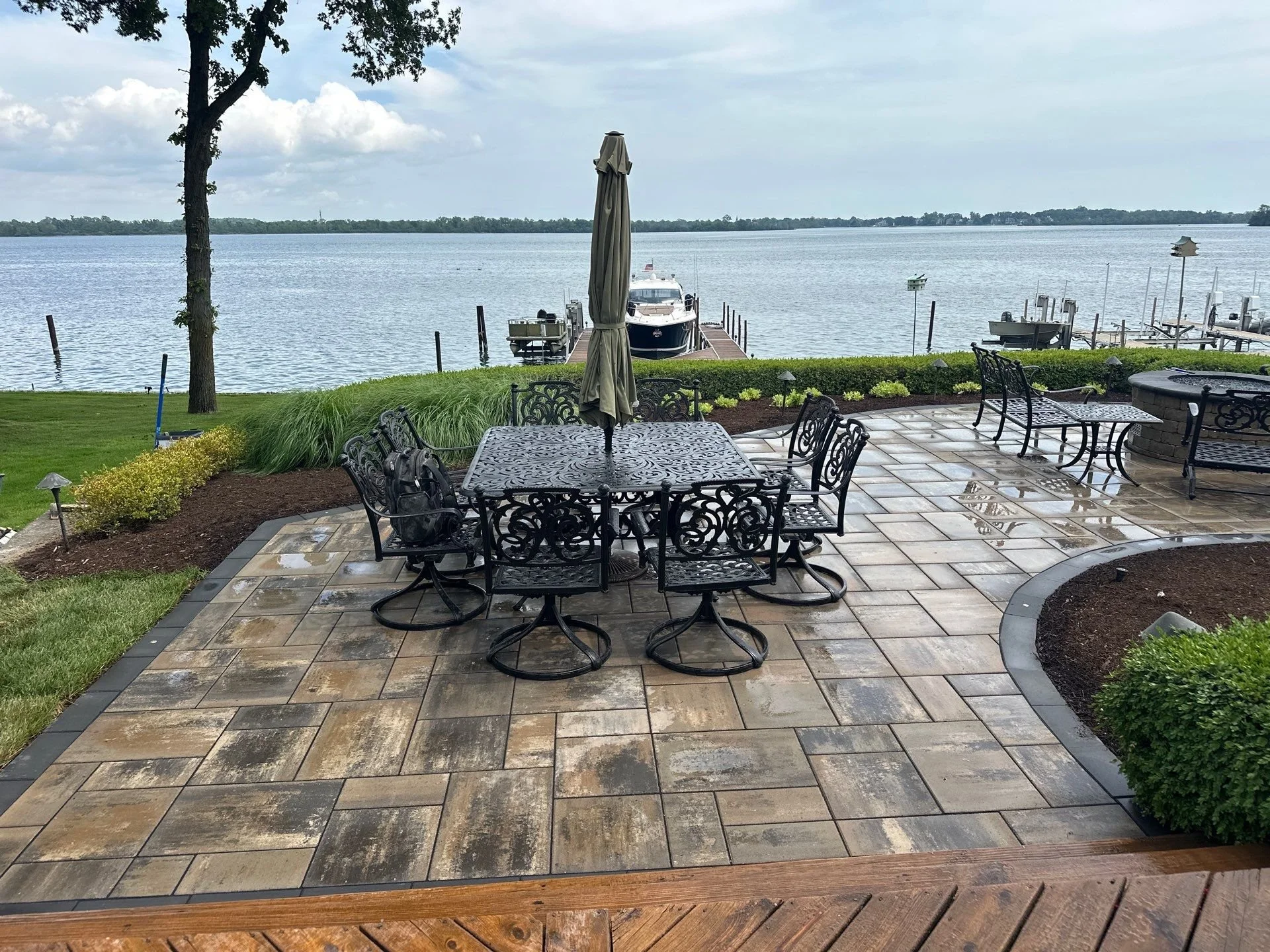 A lakeside patio with a black wrought iron dining table and chairs, a closed beige patio umbrella, and a view of a dock with boats on a body of water. The scene includes a tree, greenery, and a cloudy sky.