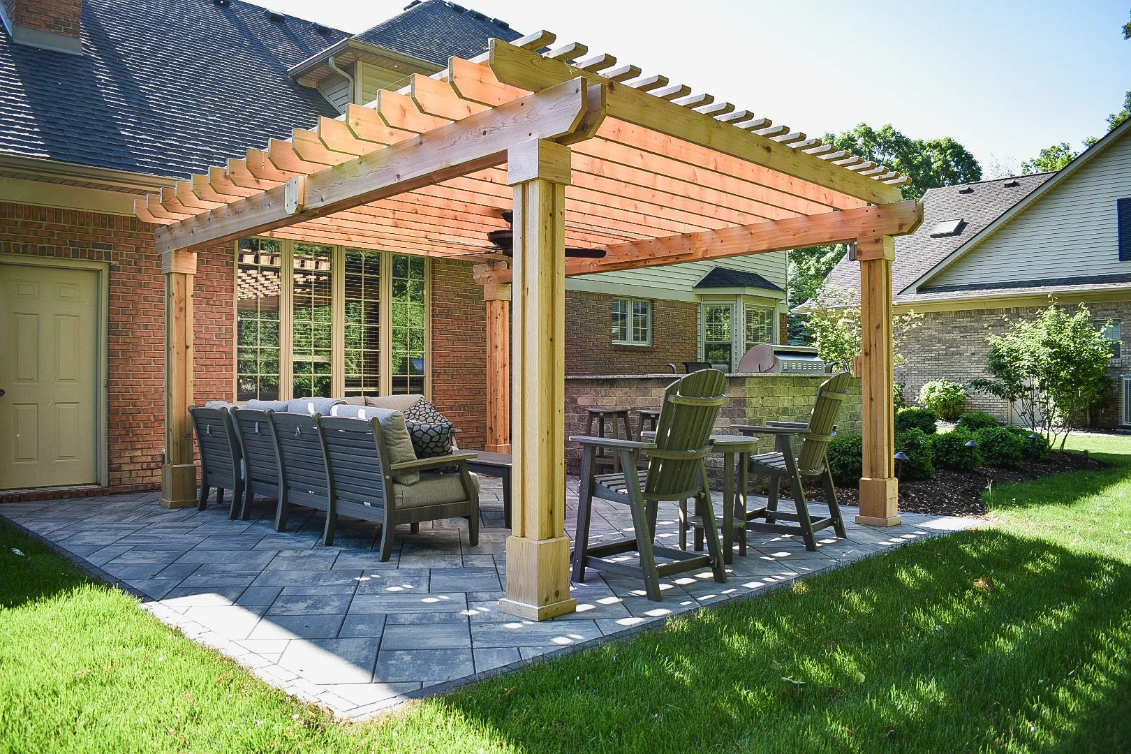 Backyard patio with wooden pergola, outdoor seating including a sofa and chairs, a grill, and landscaped lawn and bushes.