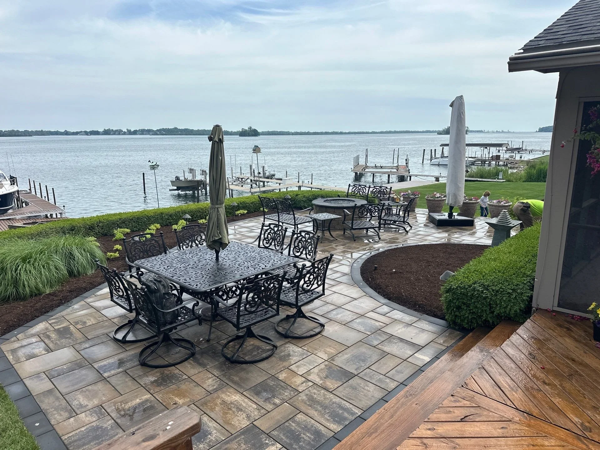 A lakeside patio with metal table and chairs, umbrellas, and potted flowers overlooking a calm lake with docks and sailboats, under a cloudy sky.