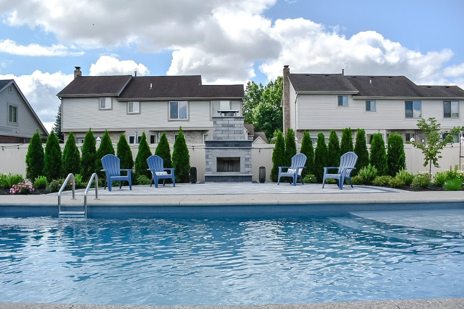 Residential backyard with a swimming pool in the foreground, blue chairs and a fireplace on the patio, green trees and shrubs, and neighboring houses with gray roofs under a partly cloudy sky — GLC Lawn & Landscape