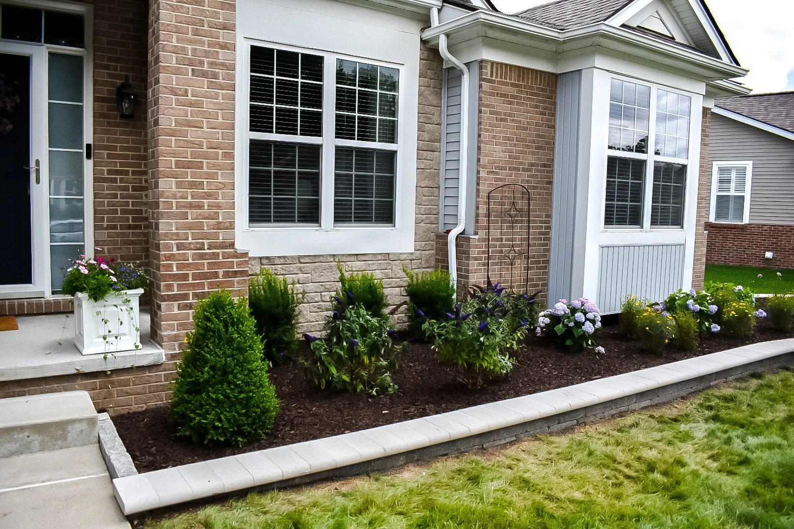 Front yard with flower bed, potted plants, shrubs, and a brick house with large white-framed windows.