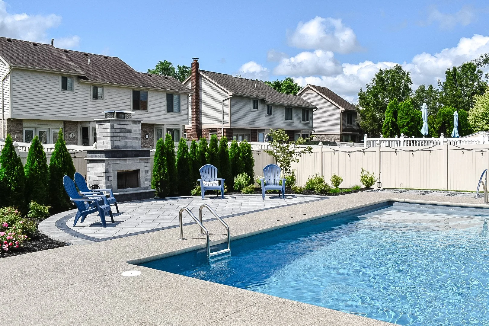 Residential backyard with swimming pool, blue chairs, a fireplace, and a white fence, with neighboring houses and trees in the background — GLC Lawn & Landscape