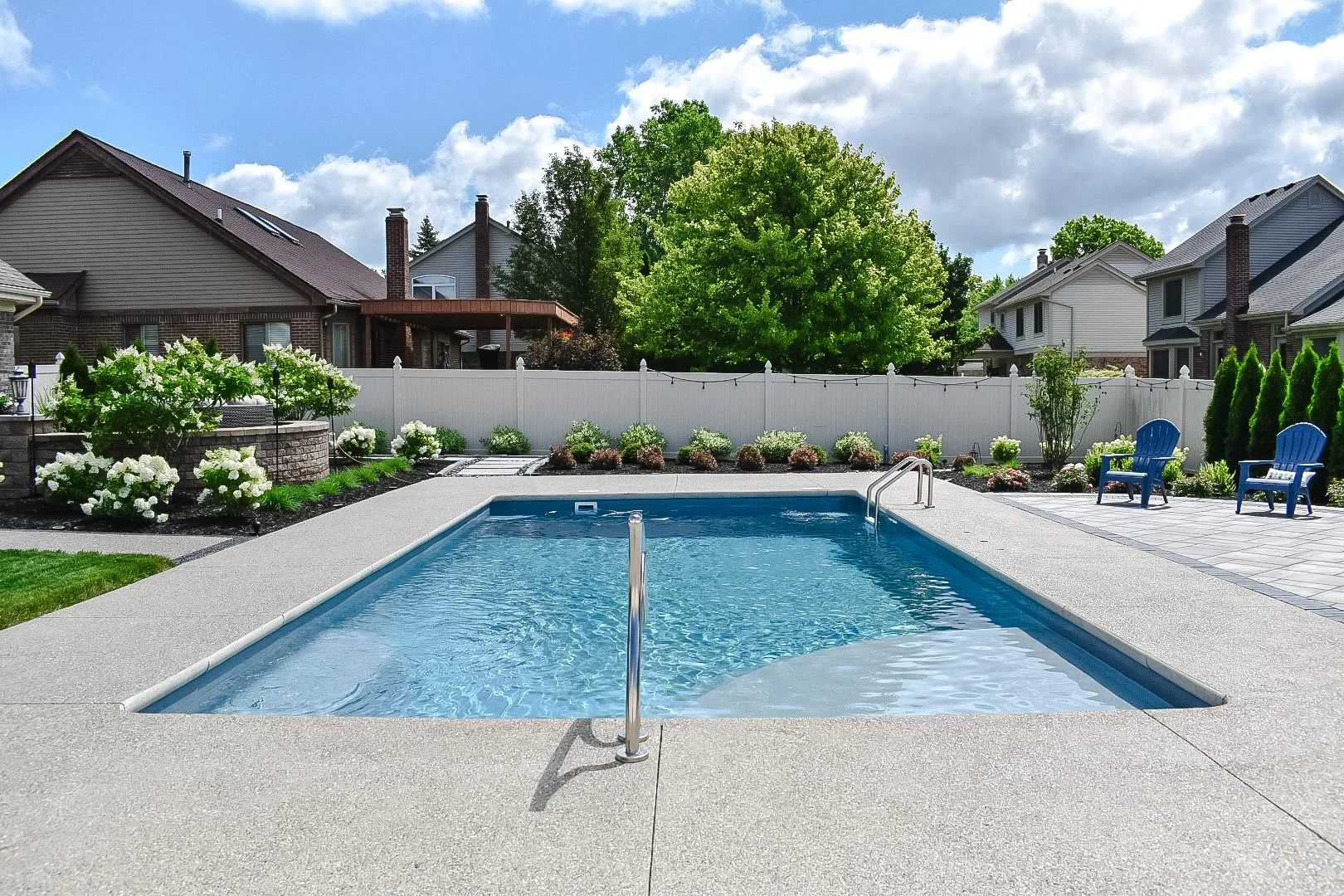 Backyard with a swimming pool, surrounded by green plants, white flowers, and two blue chairs. There is a white privacy fence and neighboring houses in the background under a partly cloudy sky.