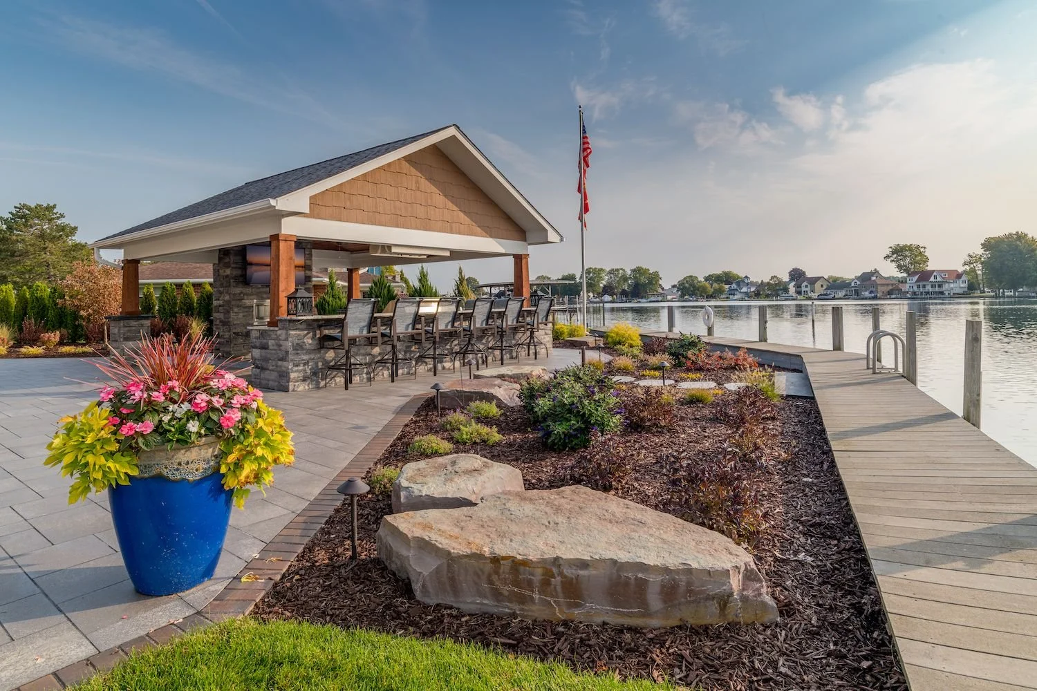 A lakeside outdoor patio with a wooden dock, a covered bar area with barstools, colorful flowers in a large blue pot, and houses across the lake, under a bright sky.