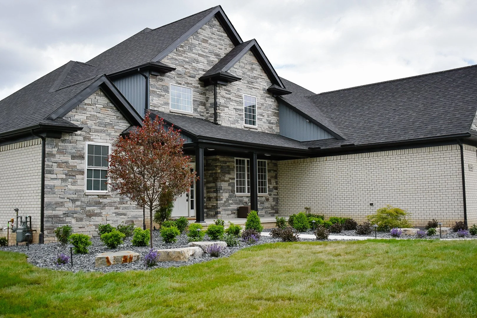 Front view of a modern house with a mix of stone and brick exterior, black roof, and landscaped garden with shrubs and a small tree.