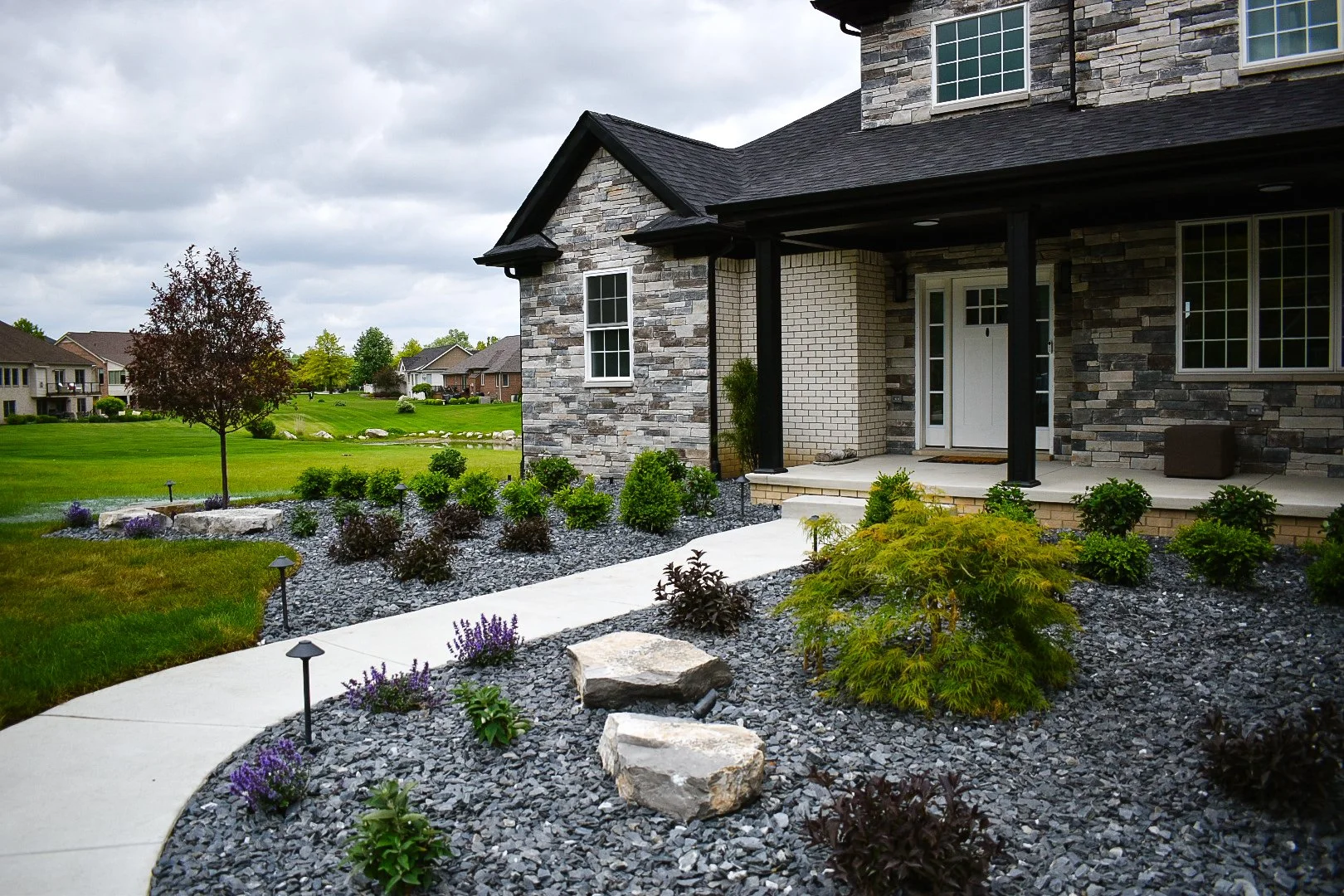Front yard of a modern house with stone and brick exterior, a concrete pathway, landscaped garden with green shrubs and purple flowers, and a tree with reddish leaves.