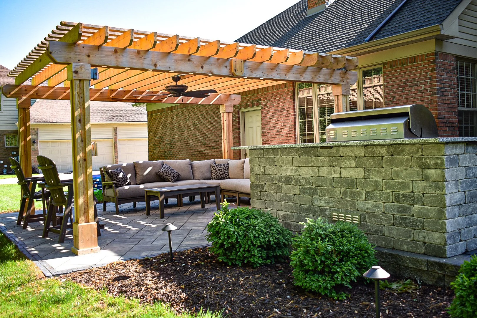 Backyard patio with a wooden pergola, outdoor sofa, chairs, brick barbecue station, and landscaped plants in front of a brick house.