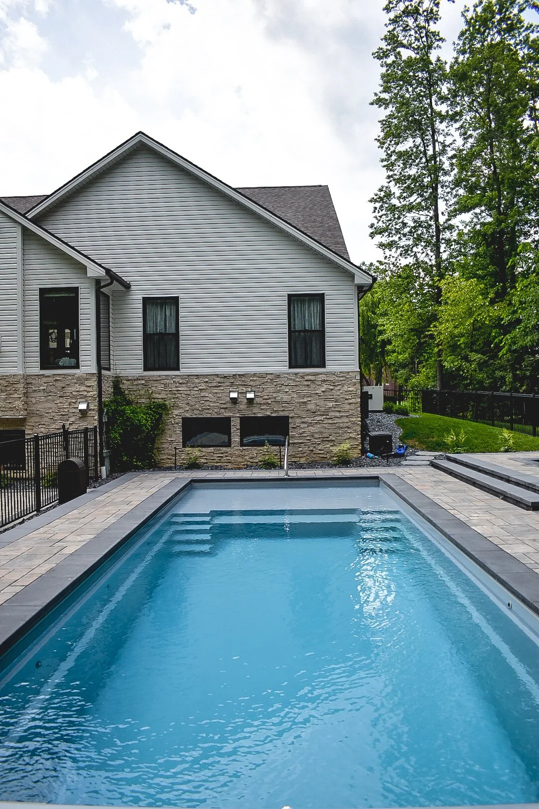 Residential backyard with a swimming pool, stone patio, surrounding fence, green grass, trees, and a white house with black windows and siding.