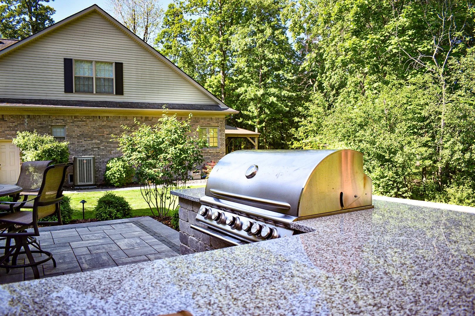 Backyard patio with grill, patio furniture, trees, and house in the background.