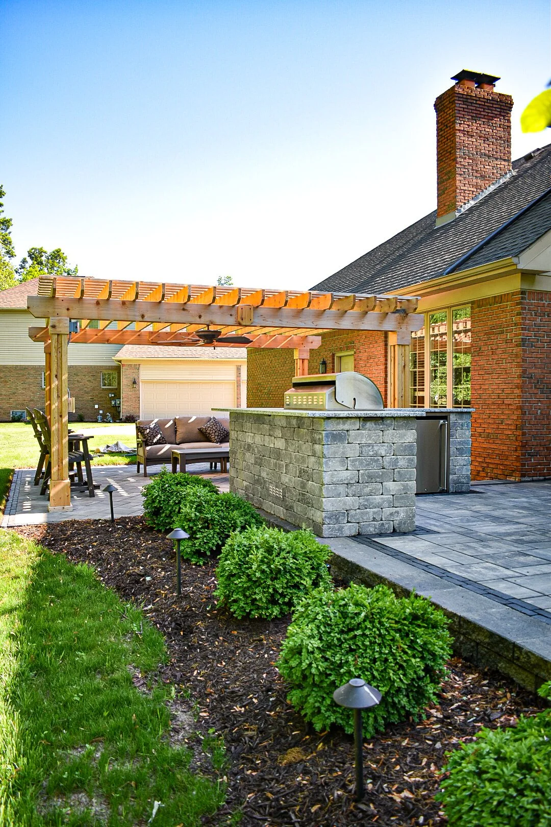 Backyard patio area with a pergola, outdoor seating, small bushes, a brick house with large windows, and a brick chimney.