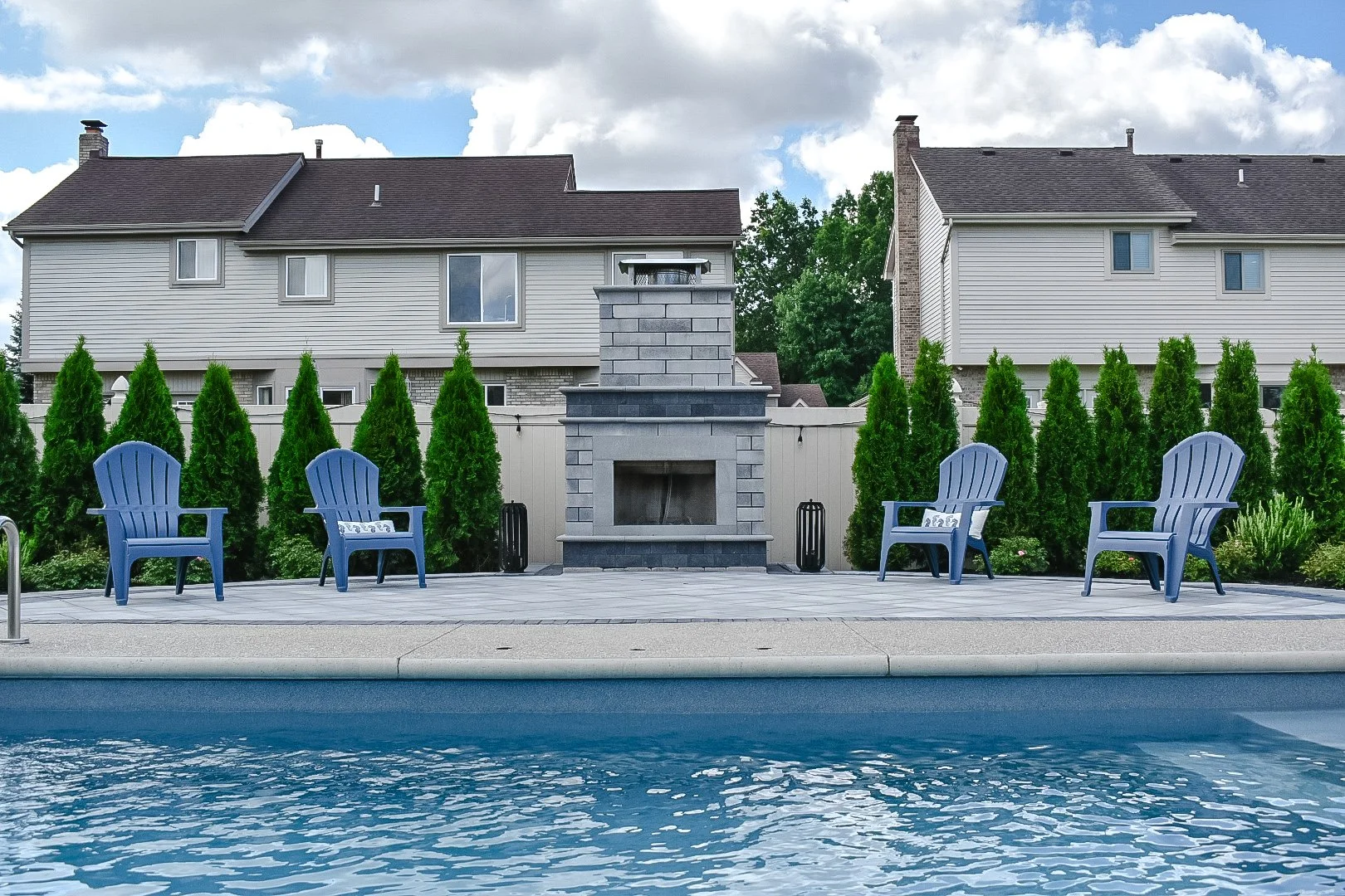 Backyard view with swimming pool, blue Adirondack chairs, a stone fireplace, and green bushes behind a white fence, with two multi-story houses in the background.