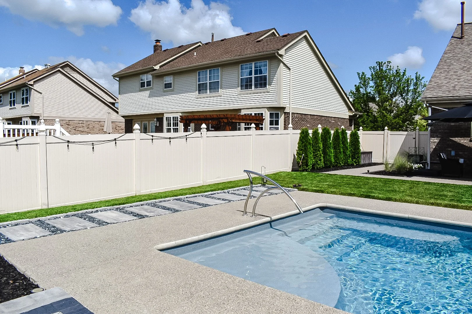 Backyard with a swimming pool, white fence, green grass, and a two-story house with siding and brick exterior under a blue sky.